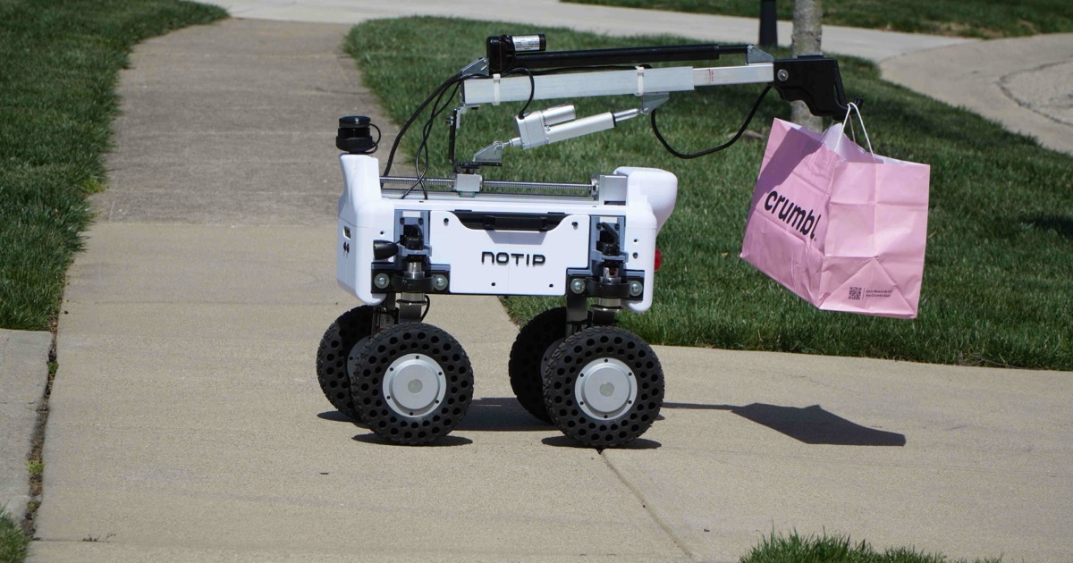 Noah, a four-wheeled white NoTip delivery robot, carrying a pink Crumbl bag up a suburban sidewalk.